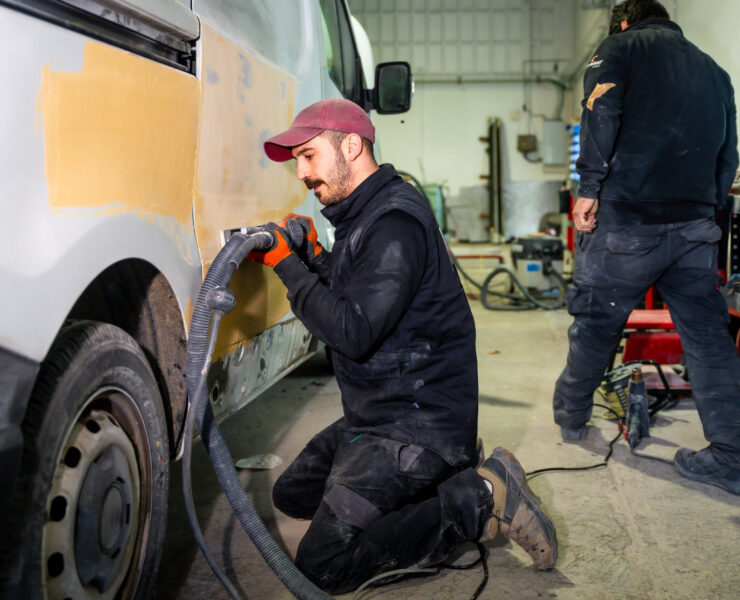 Male auto body worker kneeling and using a power sander with vacuum attachment on a van door, preparing the surface for painting in a professional repair shop alongside another technician