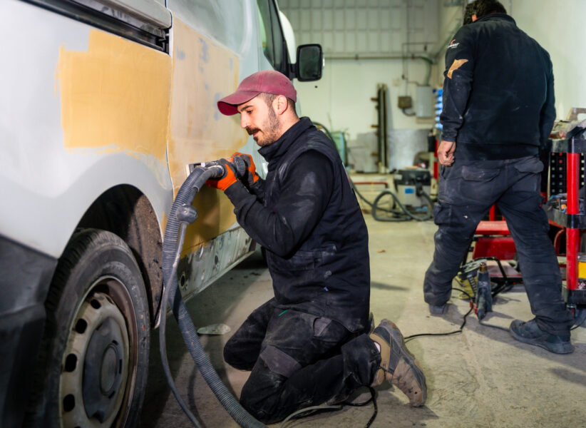 Male auto body worker kneeling and using a power sander with vacuum attachment on a van door, preparing the surface for painting in a professional repair shop alongside another technician